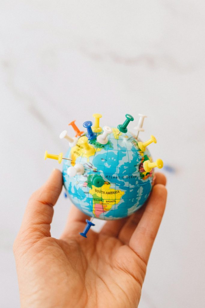 Close-up of a hand holding a mini globe with colorful push pins on a white background.